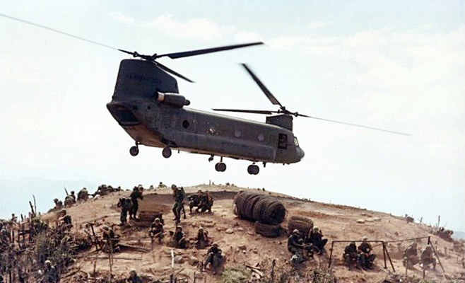Color photograph of a United States Army CH-47 Chinook helicopter, pictured in flight above a hilltop in Vietnam. There are soldiers encamped on the hilltop. The helicopter has two sets of rotors and is painted a dark grey or green. 